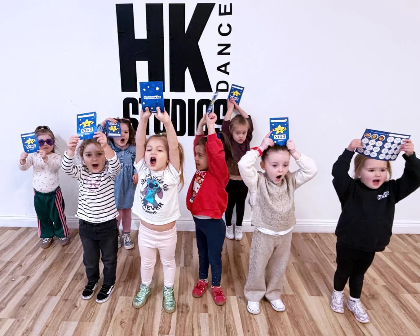 HK Tots class holding star charts overhead, mouths open mid-cheer, in front of the HK Studios studio wall.