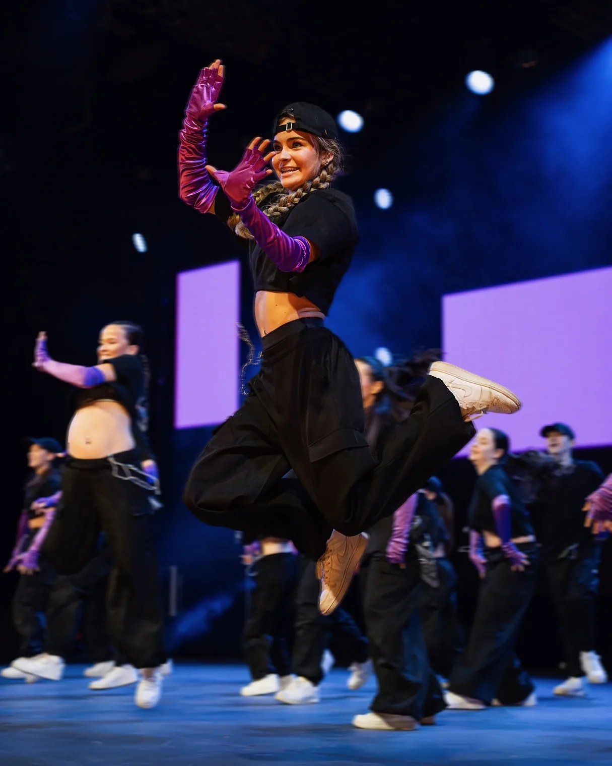 HK Dance Studios dancer mid-jump on a competition stage in black cap and purple satin gloves, ensemble in motion behind.